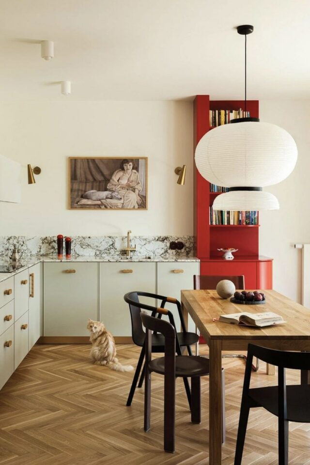 cuisine ouverte en L avec espace repas au centre table en bois meuble beige plan de travail et dosseret en marbre et bibliothèque rouge pour délimiter l'espace 