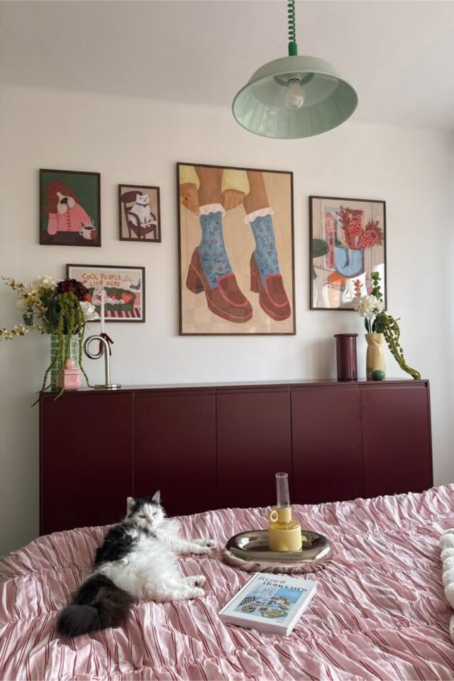commode meuble de chambre peint en couleur burgundy posée contre un mur blanc linge de lit rose pastel décoration murale tableau moderne 